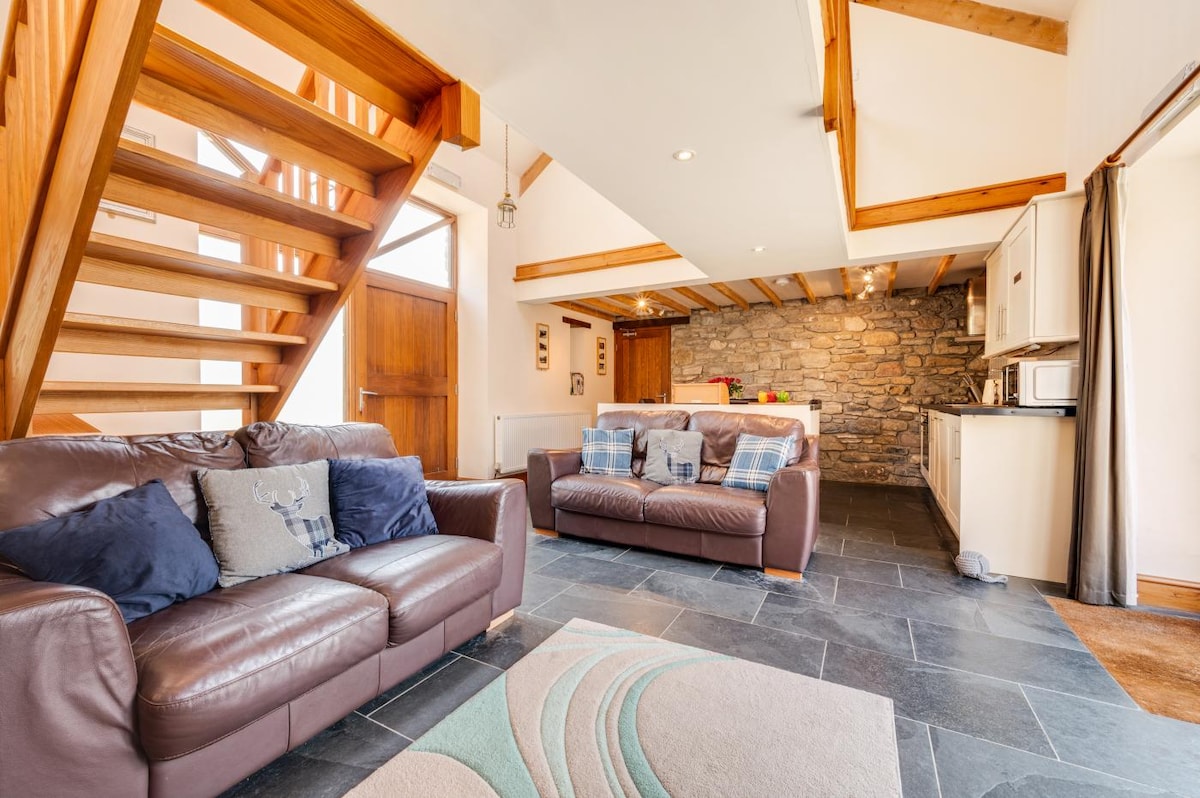 The open plan living area displays soft seating arranged for five, featuring a combination of leather sofas and decorative cushions. A wooden staircase leads to the upper floor. The space is illuminated by natural light streaming through large windows, with rustic stone walls visible in the background.