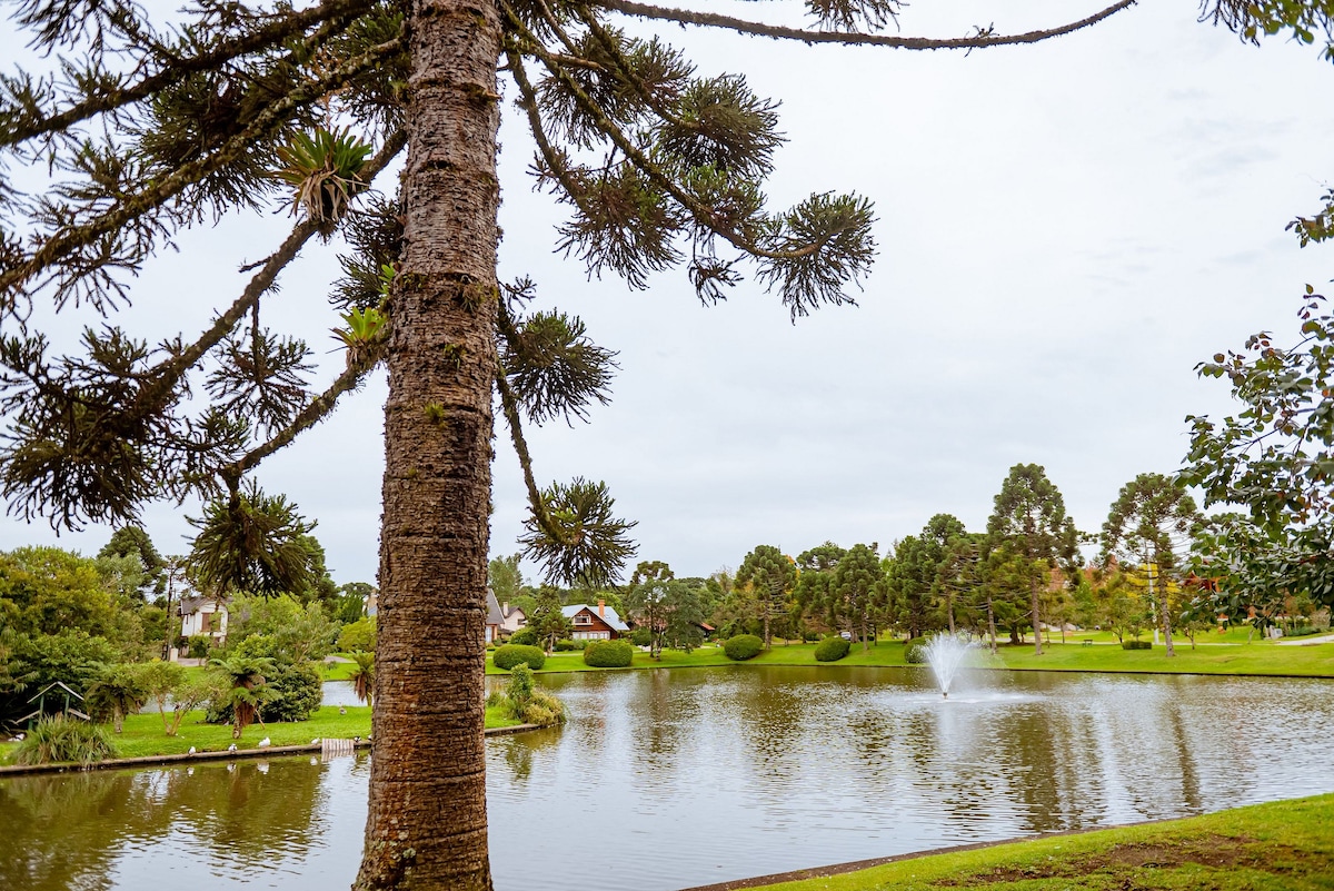 A serene lake is surrounded by lush greenery, featuring a fountain that gently sprays water. Tall trees, including a prominent araucaria, provide a natural frame to the scene, characterized by a tranquil atmosphere complemented by the idyllic landscape.