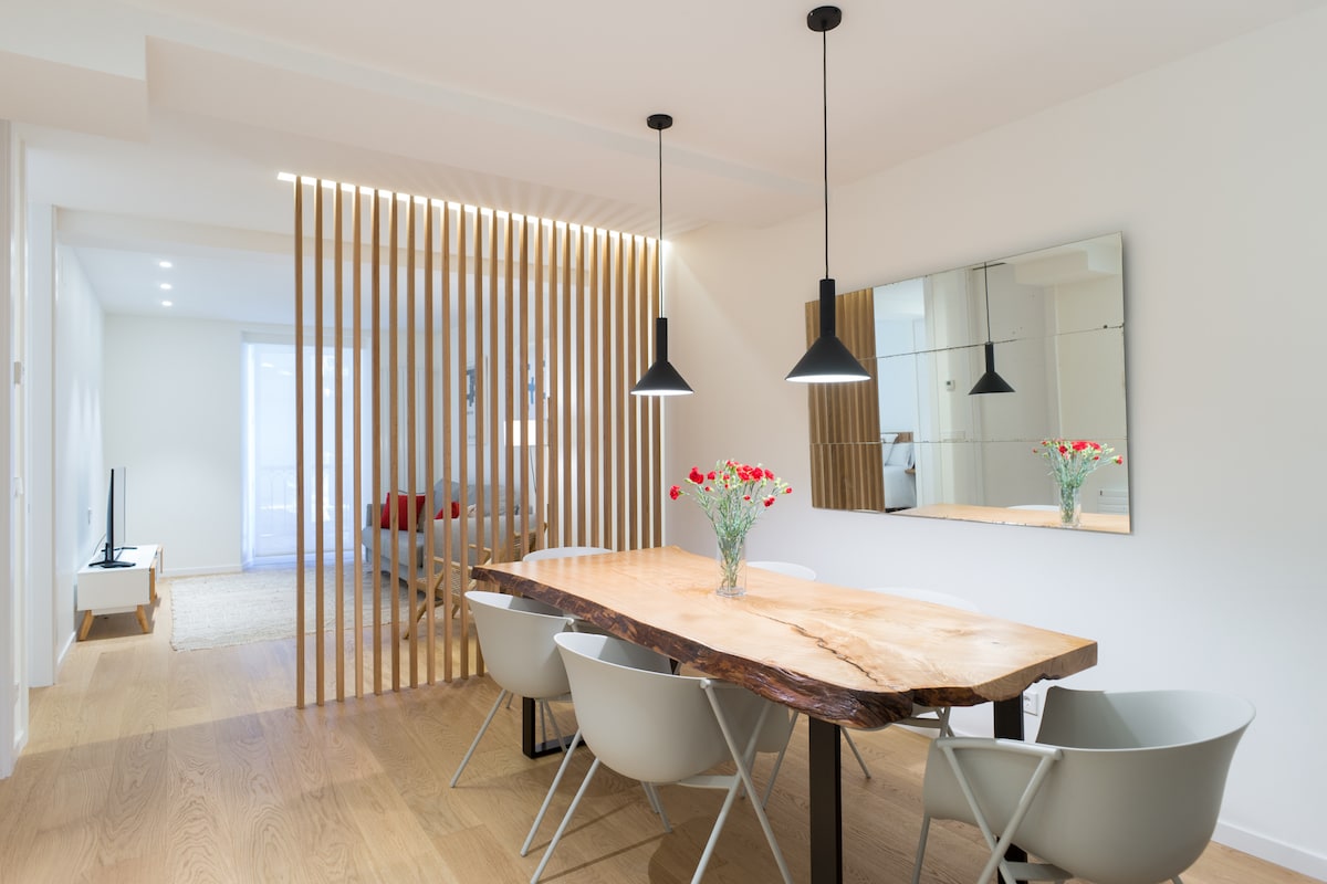 The dining area features a wooden table with a natural edge, complemented by five light-colored chairs. Above, pendant lights provide focused illumination. A mirror reflects the room’s brightness, while a small vase with flowers adds a touch of color. Behind, wooden slats create a subtle division to the living space.