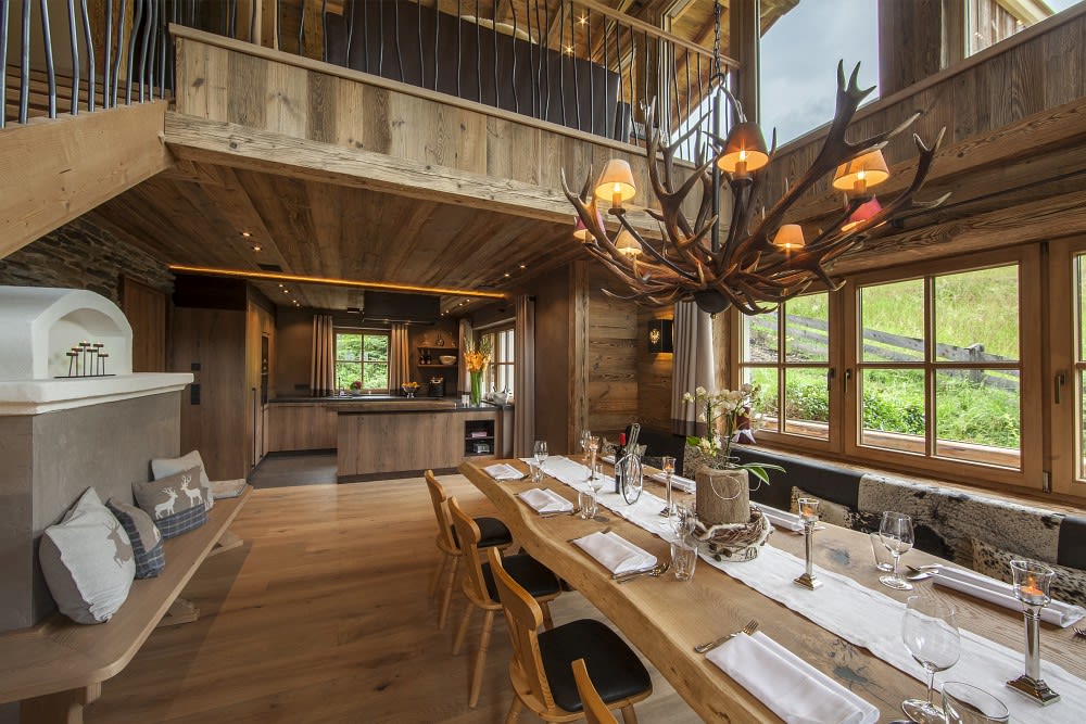 A spacious dining area features a long wooden table set for a meal, surrounded by stylish chairs. A large antler chandelier hangs above, casting warm light. The room is adorned with natural wood accents and offers views through large windows, enhancing the inviting ambiance.