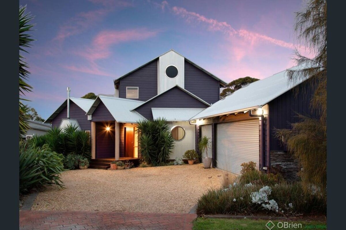 The exterior of the home is captured at dusk, showcasing a combination of dark wood and light-colored siding. Lush greenery surrounds the entrance, which features a welcoming porch illuminated by soft lighting. A gravel driveway leads to the garage, partially visible on the right.