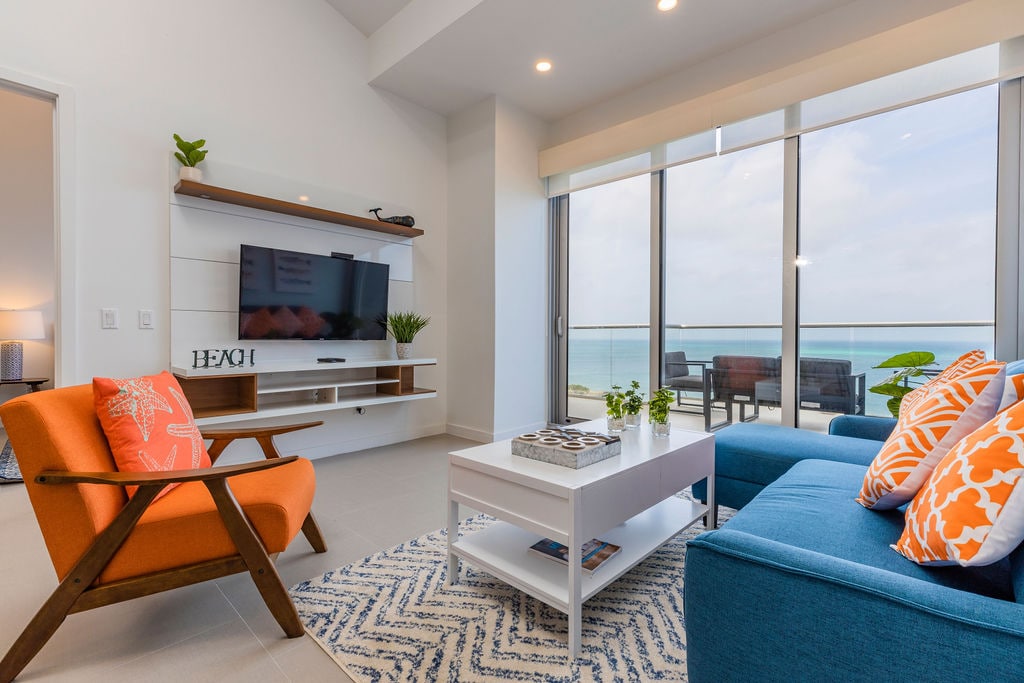 A bright living area is presented with large glass sliding doors that open to a stunning ocean view. Modern furniture includes a sofa adorned with orange and white cushions, paired with an accent chair. A white coffee table sits centrally, complemented by decorative items and a TV mounted on the wall.