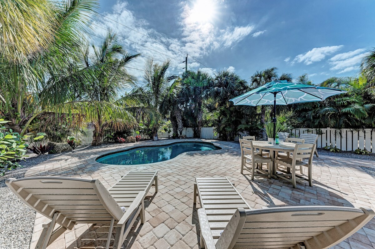 A spacious outdoor area features a heated lagoon-style pool surrounded by tropical landscaping. Two lounge chairs are positioned near the pool, complemented by a dining table set with an umbrella for shade. The sun casts a bright light over the scene, enhancing the inviting atmosphere.