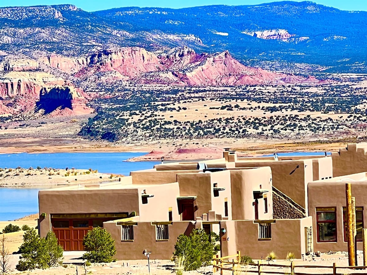 The Grand Hacienda Inn is set against a backdrop of rolling hills and dramatic cliffs, overlooking Abiquiu Lake. The earthy exterior reflects traditional adobe architecture, complemented by lush greenery around the property. The expansive vista showcases vibrant colors of the landscape under clear blue skies.