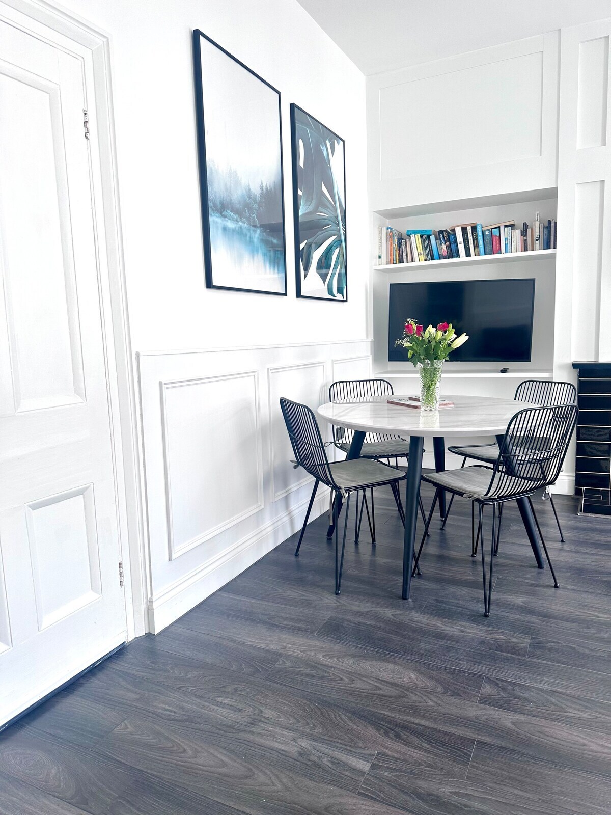 A dining area features a round table surrounded by four black wire chairs. Two framed artworks adorn the white walls, and a mounted television sits above a shelf filled with books. The dark wood flooring adds warmth to the space.