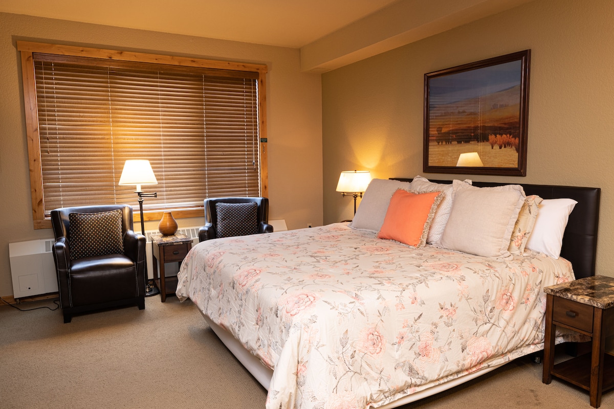 The room features a spacious bed with a floral patterned quilt and multiple pillows. Two club chairs and a side table are positioned beside the bed. Soft lighting from table lamps provides a warm glow. A large window adorned with wooden blinds allows natural light to enter.
