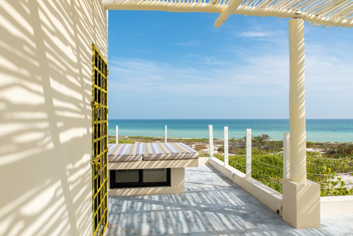 A spacious terrace features a lounging bed with striped cushions, providing an unobstructed view of the ocean. Sunlight casts intricate shadows across the surface, while the calming blue water stretches to the horizon, framed by gentle coastal vegetation.