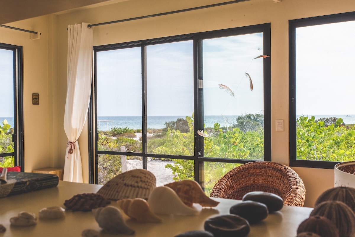 Large windows provide views of the serene ocean and lush coastal vegetation. Natural light floods the interior, which features simple decorative elements such as seashells and smooth stones. The space is designed to connect guests with the surrounding beauty of nature.