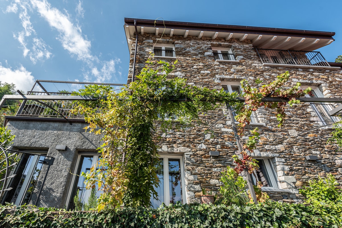 The stone exterior of the building reflects rustic architecture, complemented by vibrant green climbing plants. Large windows and multiple balconies enhance the aesthetic, providing views of the surrounding nature and sky.