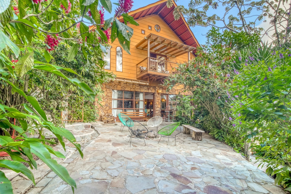 A charming outdoor seating area features two stylish chairs set on a stone patio, surrounded by lush greenery and colorful flowers. The wooden structure of the house is visible in the background, showcasing a balcony and large windows that invite natural light.