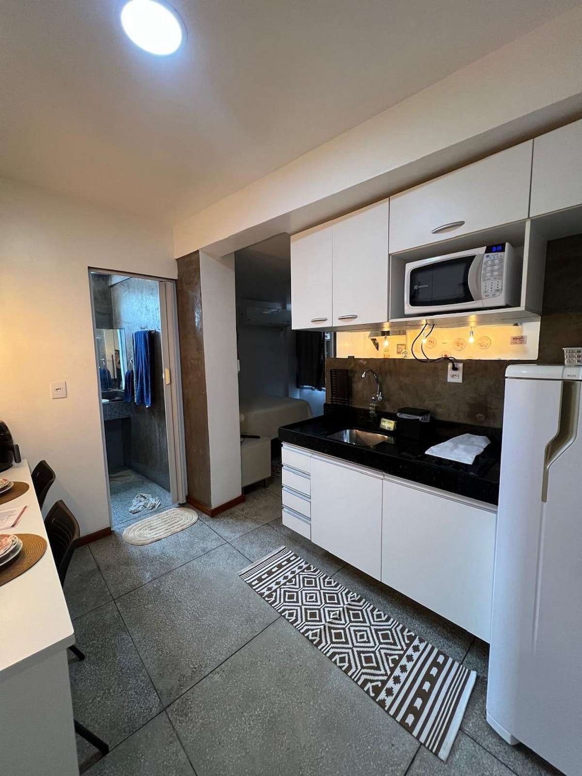 A compact kitchen features modern white cabinetry and a dark countertop. Appliances include a microwave and a refrigerator. A small dining area with black chairs is visible, along with a patterned rug on the floor. A doorway leads to another space, enhancing the layout.