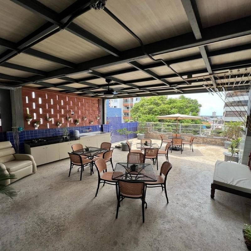 A spacious common area is highlighted by a relaxed seating arrangement, featuring multiple tables and chairs. A kitchenette is visible along one wall, with greenery adding a touch of nature. Large windows open up to a city view, allowing natural light to flood the space.