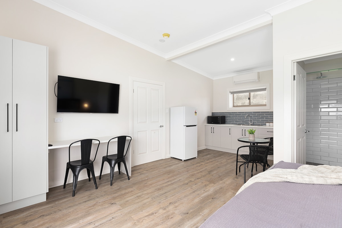 A bright, open living space features a queen bed with a soft grey blanket. A kitchenette with a fridge and sink is visible, alongside a wall-mounted TV. Two black chairs are positioned at a small table, and a window provides natural light, illuminating the wooden floor.