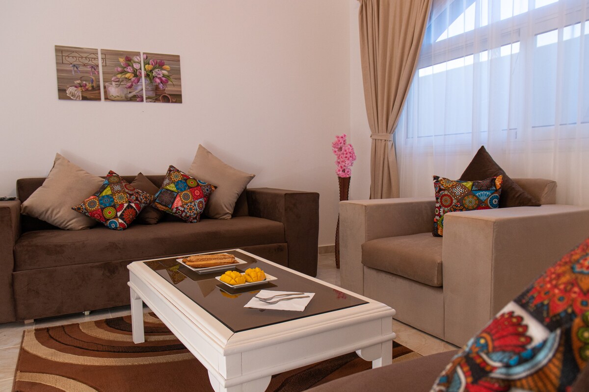 A cozy living area is furnished with two sofas adorned with decorative pillows and a coffee table. A tray with snacks is displayed on the table, while light filters through sheer curtains, creating a serene ambiance. Wall art featuring floral designs adds a touch of color.