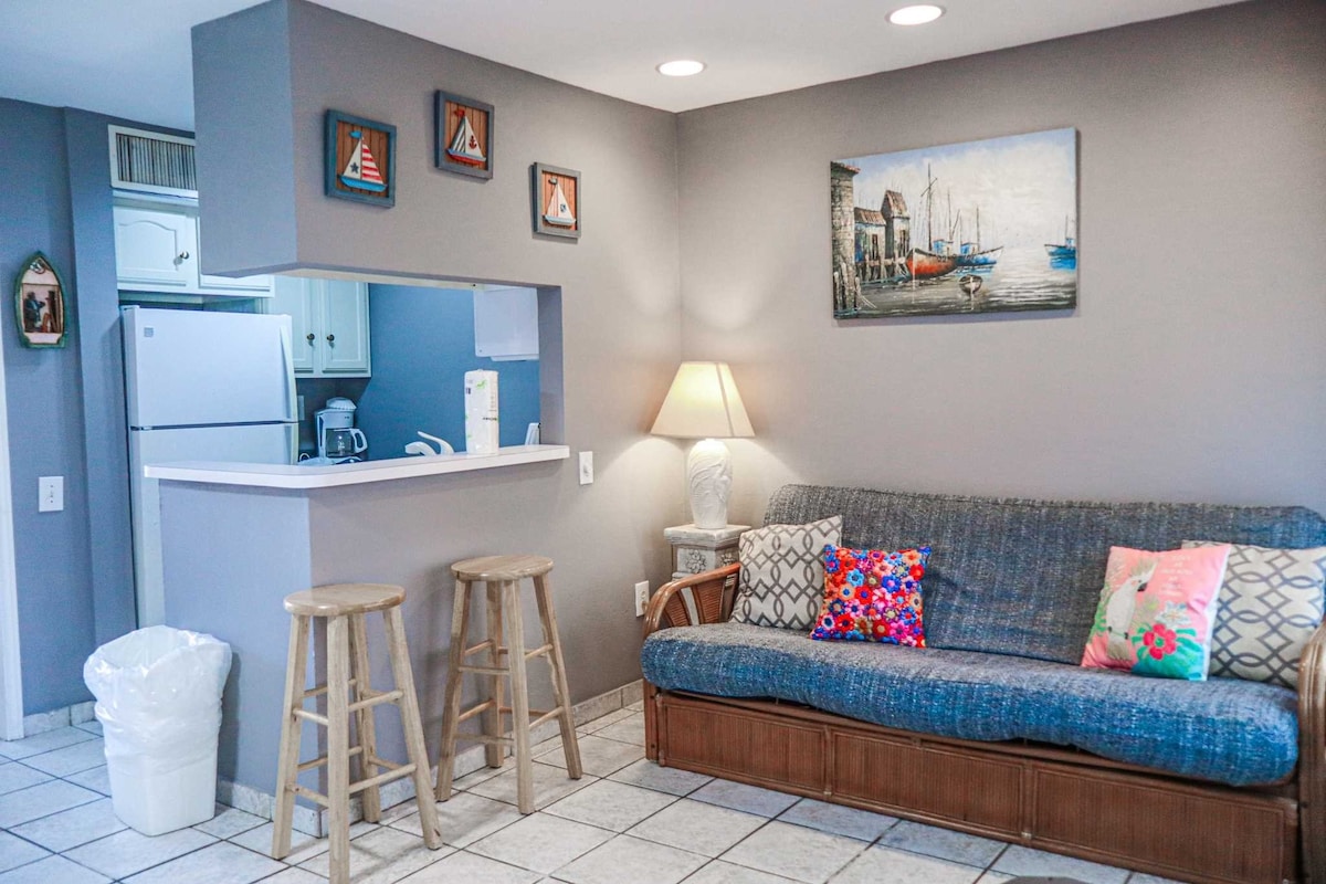 A cozy living area features a blue futon with colorful throw pillows perched against a light gray wall. Above, a painting of a harbor scene is displayed. Two wooden stools are positioned near a service counter, and a lamp provides soft lighting. A kitchen area is visible in the background.