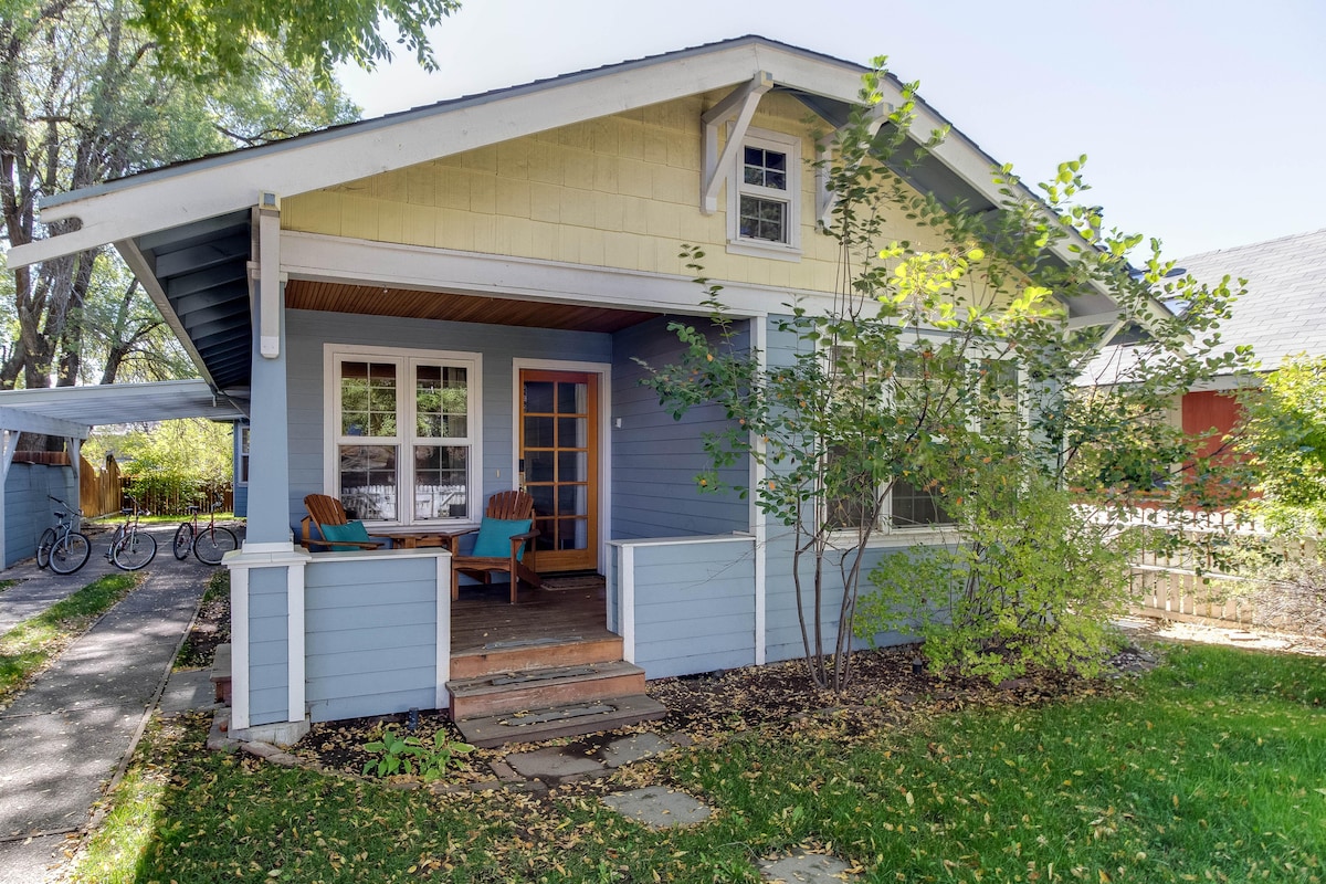 The exterior of a charming bungalow is showcased, with a light blue facade and inviting porch. Two Adirondack chairs are placed on the porch, surrounded by greenery. A pathway leads to the front steps, and bicycles can be seen parked nearby.