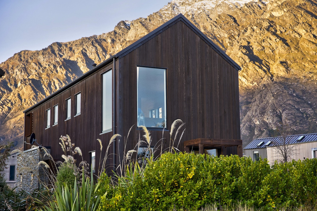 The exterior of a dark cedar house is showcased against a backdrop of rugged mountains. Large, modern windows reflect natural light, while surrounding greenery adds to the serene environment. The home’s unique architecture harmoniously integrates with the picturesque landscape.