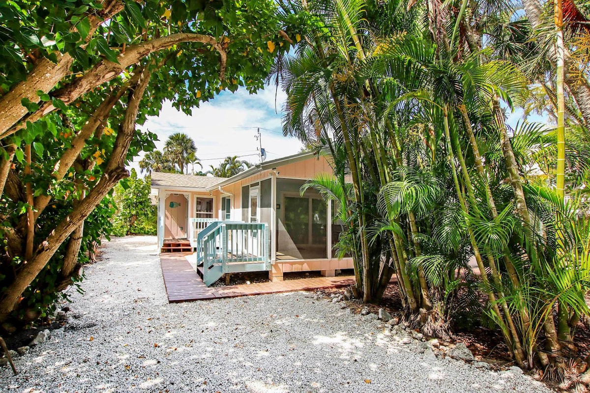 A charming cottage is seen nestled among tropical greenery, accessed by a gravel pathway. The front porch features a wooden railing and steps leading to a door, creating an inviting entry. Lush palms and vibrant foliage surround the space, enhancing the secluded atmosphere.