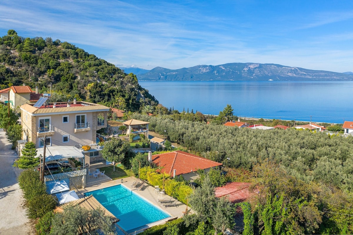 An aerial view captures the mansion surrounded by lush greenery and gardens, with the large swimming pool prominently placed. Nearby structures and the serene sea stretch into the distance, framed by hills under a clear blue sky.