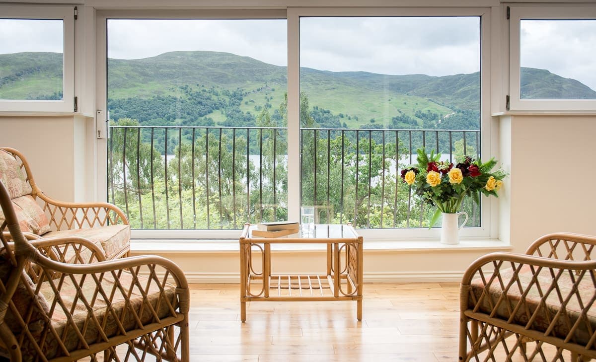 Large windows offer sweeping views of the lush green hills and Loch Rannoch. A small coffee table sits between two wicker chairs, while a vase of colorful flowers adds a touch of nature indoors. The light hardwood floor complements the serene atmosphere.