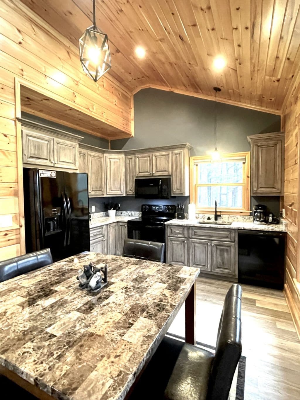 A modern kitchen features wooden cabinetry and a granite countertop. Stainless steel appliances are present, including a refrigerator and oven. A dining table with black leather chairs occupies the center, while natural light filters through a window, enhancing the warm wood accents.