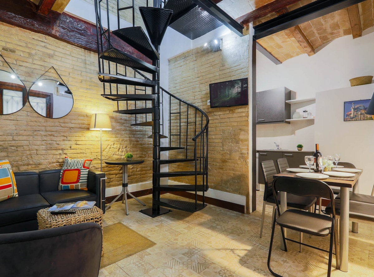 A spiral staircase leads to the upper level of a duplex, with exposed brick walls adding texture. A seating area with a dark sofa, decorative pillows, and a side table is complemented by a dining space featuring a table set for four. Soft lighting creates a cozy ambiance.