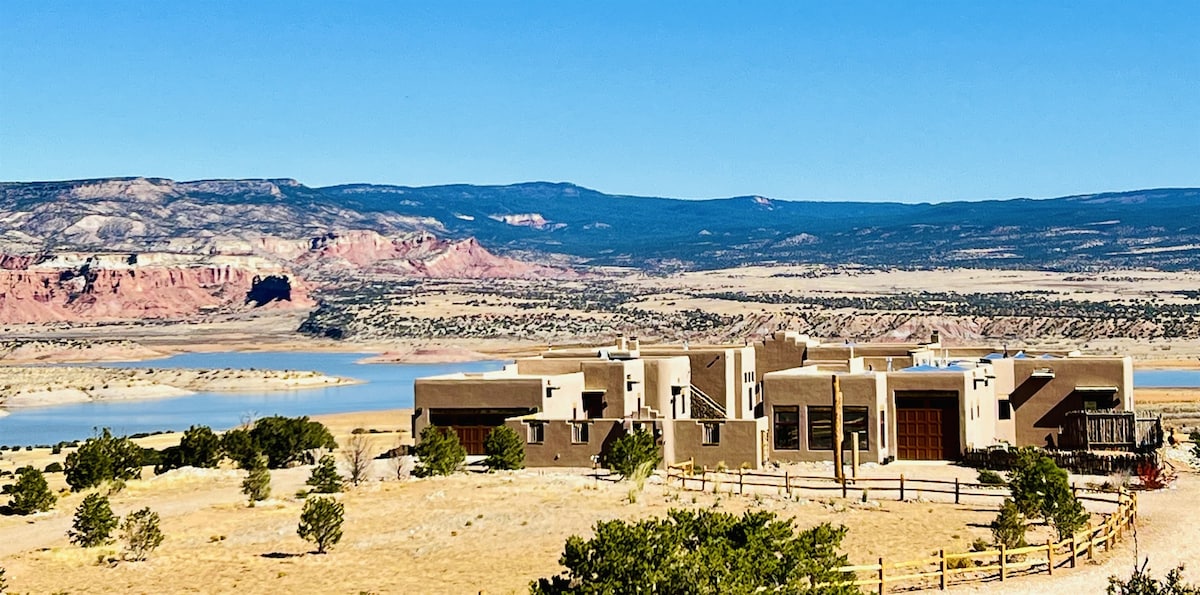 A spacious adobe-style building is set against a backdrop of expansive landscapes, featuring rolling hills and Abiquiu Lake shimmering in the sunlight. Dramatic cliffs with vibrant hues rise in the distance, highlighting the natural beauty of the surroundings.