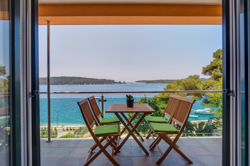 A balcony view is presented, featuring a wooden table surrounded by four folding chairs with green cushions. The scene overlooks the sea, showcasing a calm blue water landscape, framed by lush trees along the shore.