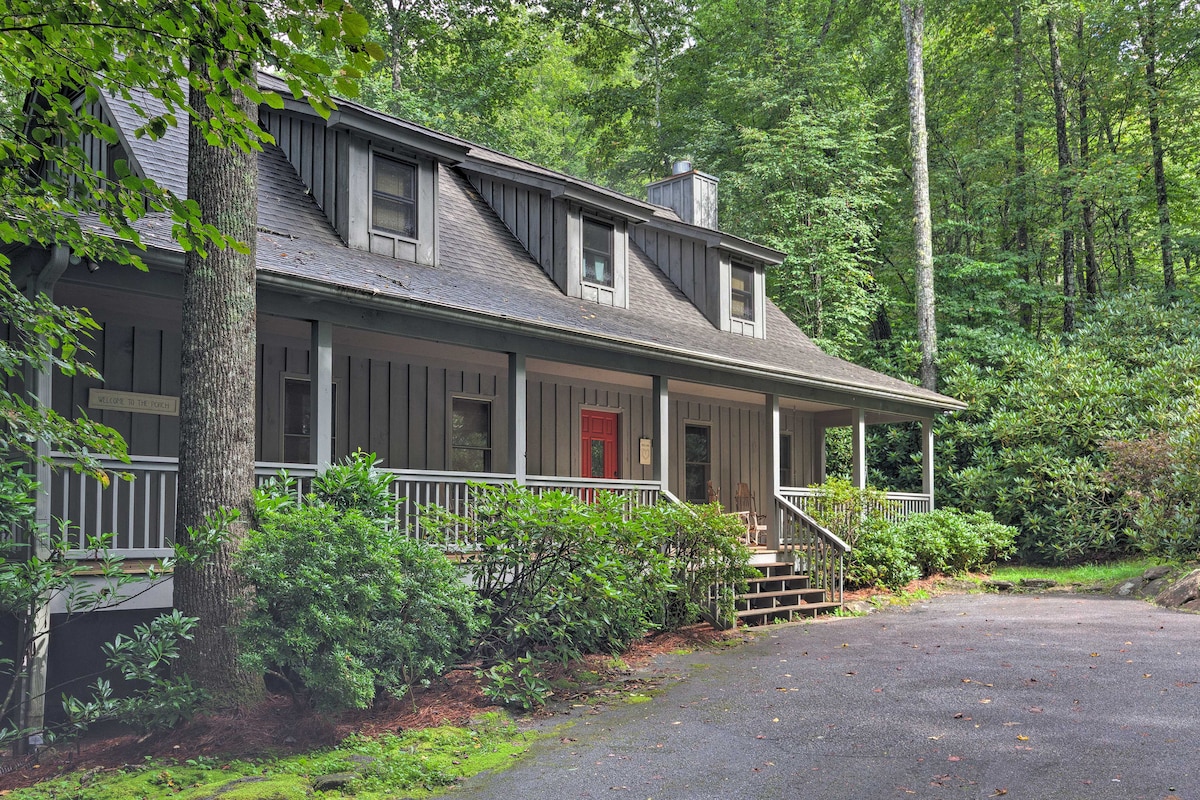The exterior of the cabin showcases a staggered roofline with a welcoming front porch bordered by greenery. A staircase leads to the entrance, flanked by flowering shrubs and trees, offering a serene woodland setting.