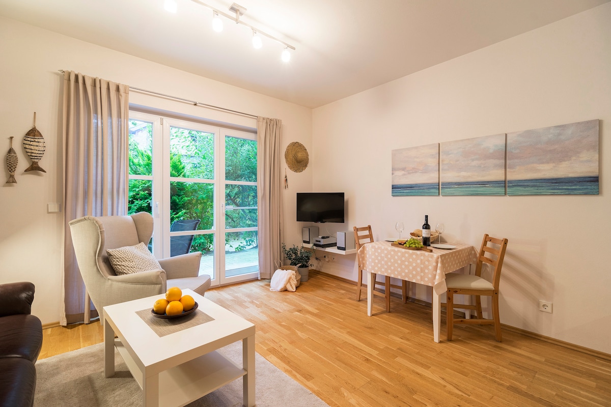 A bright living area features a light-colored sofa and a dining table set for four. Large glass sliding doors open to a private terrace, allowing natural light to fill the space. Wall art depicting serene coastal scenes adds a subtle decorative touch.