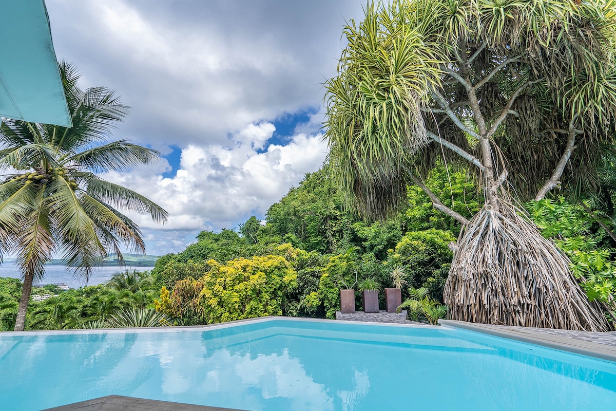 An infinity pool features a clear blue surface reflecting the sky, bordered by lush greenery. A tall Pandanus tree provides shade nearby, while two dark lounge chairs are positioned near the pool's edge, inviting relaxation amidst the tropical landscape.