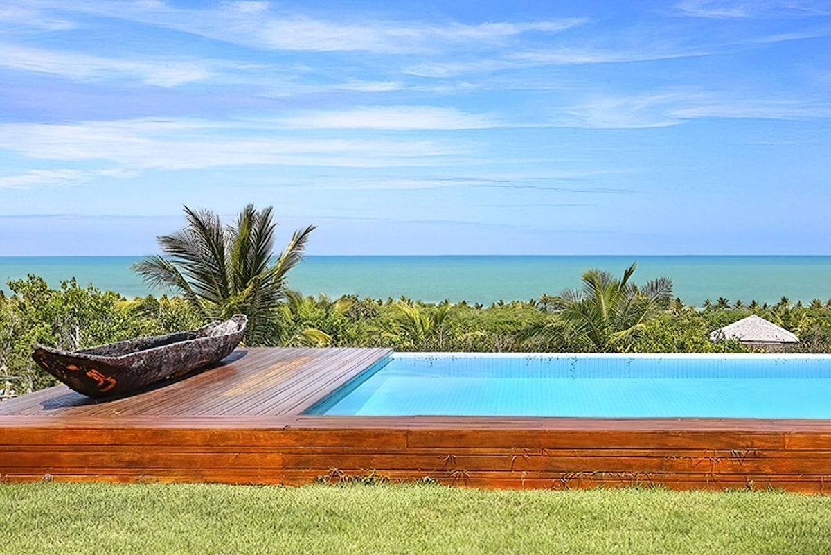 An infinity pool extends towards the horizon, merging with the distant sea beyond. Lush tropical vegetation surrounds the area, with palm trees framing the view. A rustic wooden canoe rests on the pool deck, adding a natural element to the serene landscape.