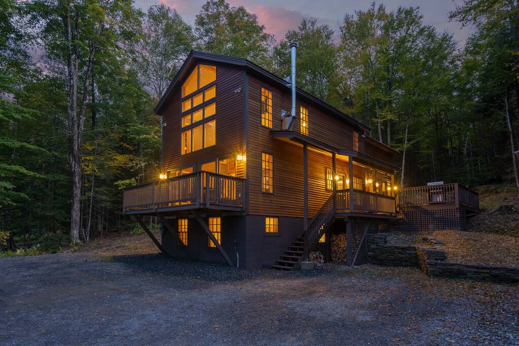 A modern wooden house is positioned in a wooded area, showcasing multiple large windows that emit a warm glow. A sizable deck wraps around the upper level, offering ample space for seating. The structure is elevated, with a stone staircase leading to the entry.