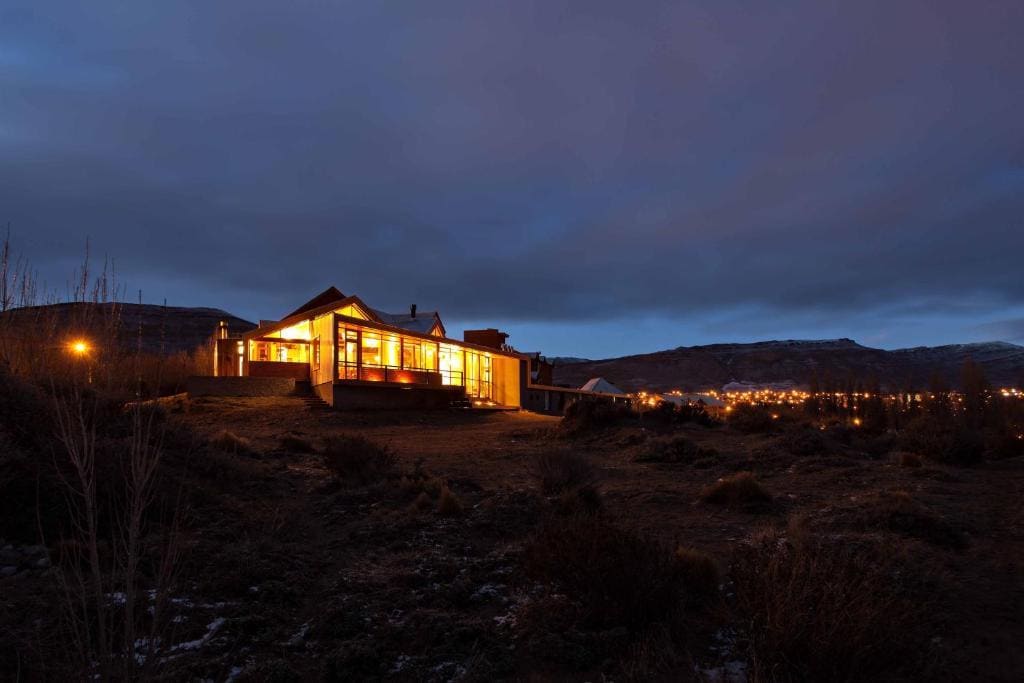 A modern Patagonian-style building emerges against a twilight sky, illuminated warmly from within. The expansive windows showcase the interior glow. In the background, the softly lit city of El Calafate rests beneath the shadow of the mountains.