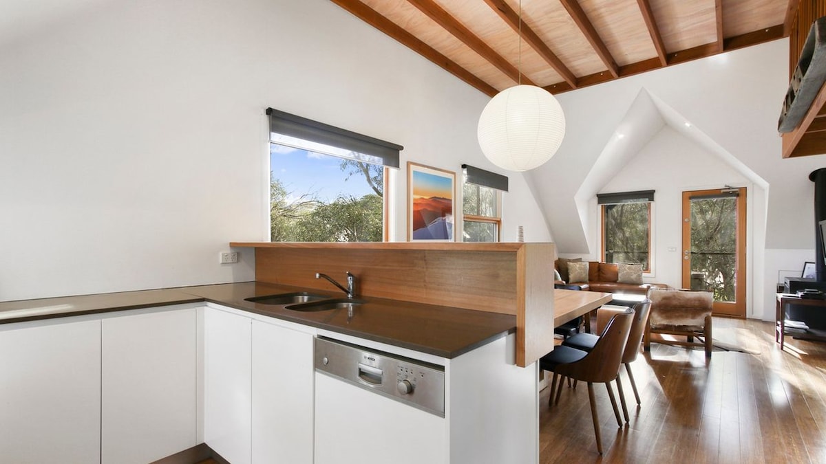 An open plan living area features a modern kitchen with white cabinetry and a dark countertop. Natural light enters through large windows, highlighting the dining area with a wooden table and dark chairs. The ceiling showcases exposed beams, contributing to a spacious atmosphere.