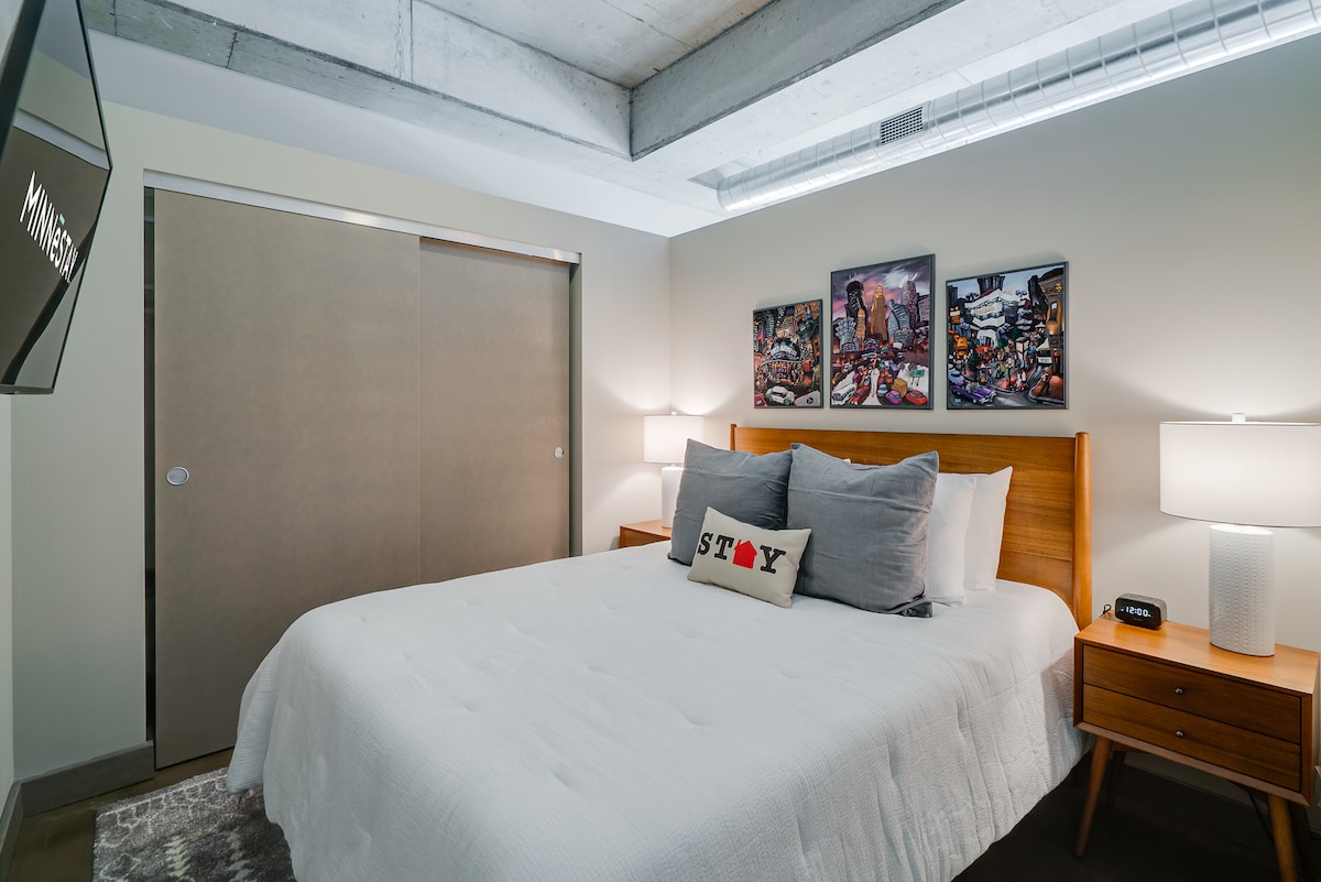 A cozy sleeping area is presented with a spacious bed dressed in crisp white bedding. Flanking the bed are two stylish nightstands, each topped with a lamp. Colorful artwork from local artists is displayed on the wall behind the bed, enhancing the modern design.