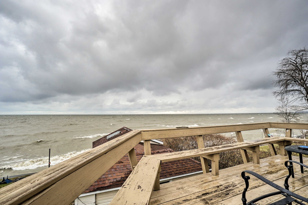 A view from the wraparound deck showcases a scenic expanse of Lake Erie, with gray clouds overhead and gentle waves lapping at the shore. The deck provides a spacious area to relax, framed by the cottage's wooden railing.