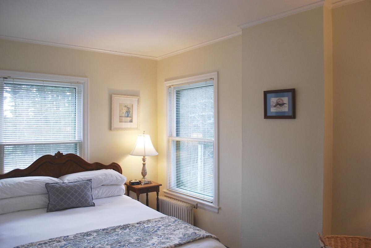 A cozy guest room features soft yellow walls and natural light filtering through two windows with blinds. A queen bed is adorned with a patterned blue and white throw. A nightstand with a lamp is positioned beside the bed, creating a warm ambiance.