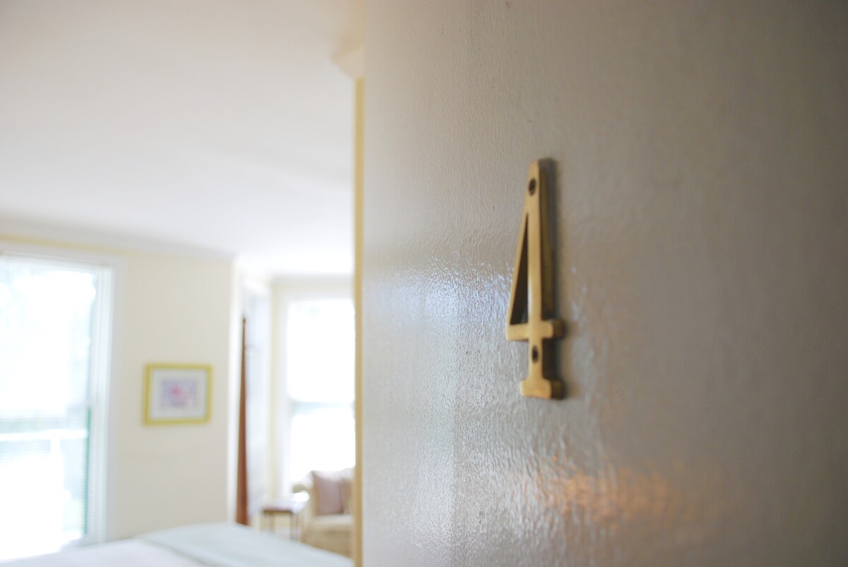 The number 4 is displayed in a subtle brass finish on a light-colored wall. Soft natural light enters through nearby windows, illuminating the serene atmosphere of the guest room entrance.