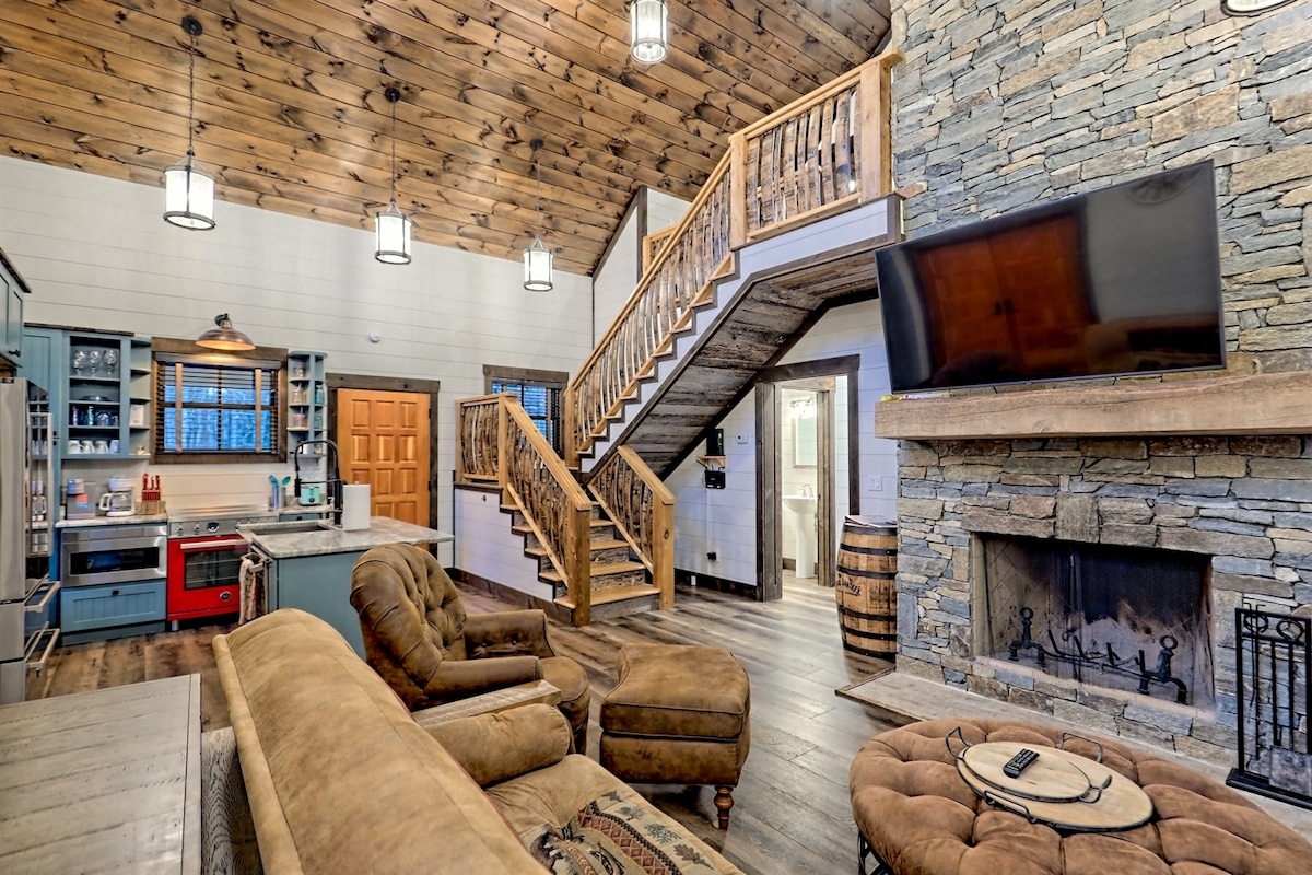 The living area showcases a grand stone fireplace beneath a high wooden ceiling. A large sectional sofa faces a mounted television. A whiskey barrel and a staircase with wooden railings create visual interest, while the open kitchen features cabinets and appliances that complement the rustic design.