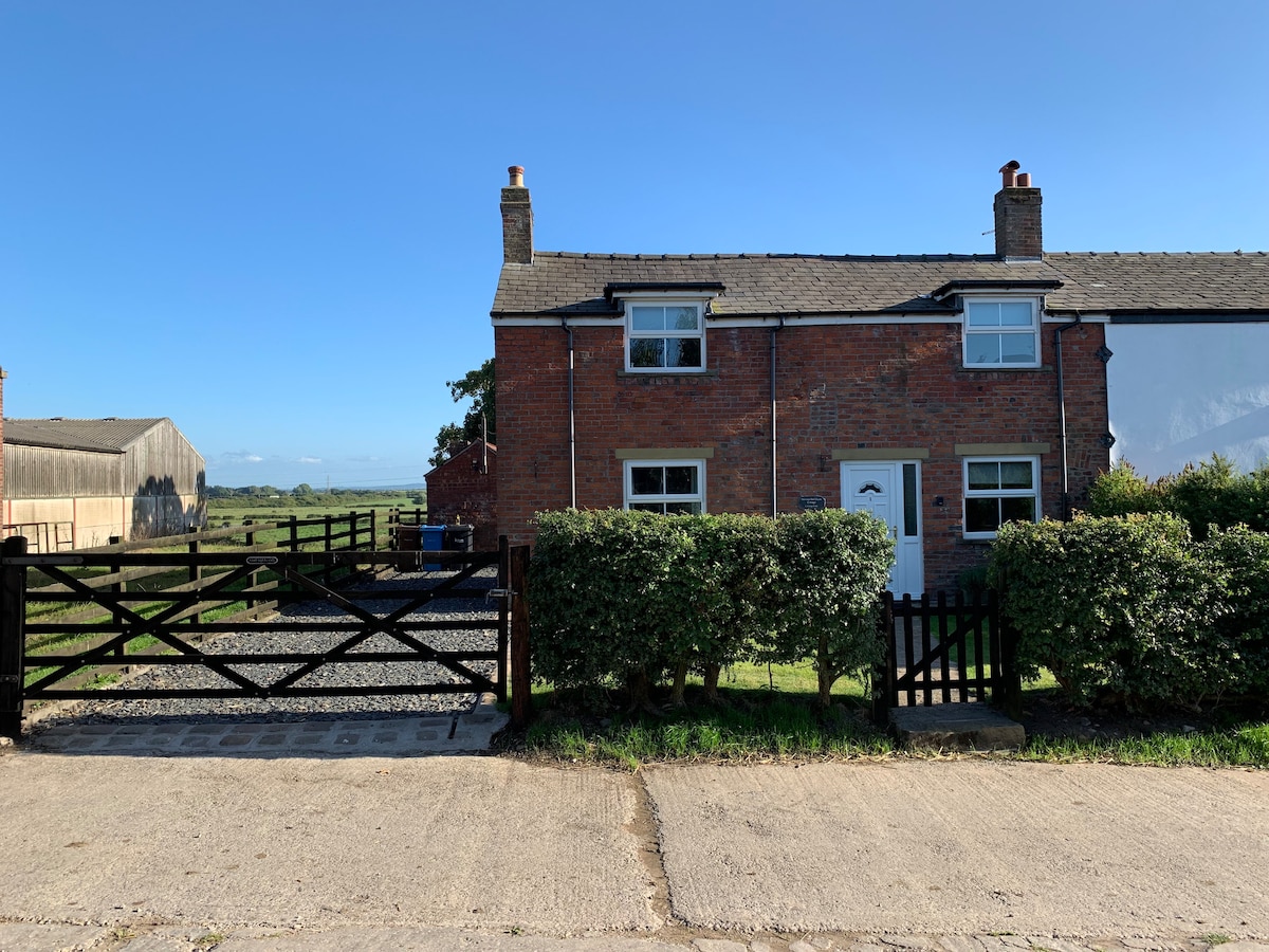 A charming cottage is framed by neatly trimmed hedges and a gated gravel drive. The brick exterior features traditional windows and a welcoming front door. Agricultural buildings are visible in the background, set against a clear blue sky.