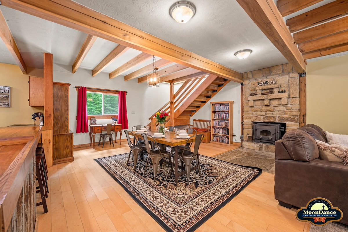 A spacious living area features a large dining table surrounded by chairs, complemented by a comfortable sofa. Wooden beams create a warm atmosphere, while a stone fireplace adds a rustic touch. Natural light fills the room, enhancing the inviting decor.