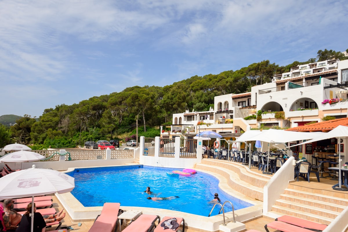 A shared outdoor area features a rectangular swimming pool surrounded by sun loungers. Umbrellas provide shade over tables and chairs, while lush greenery forms the backdrop. Guests can be seen enjoying the pool, creating a relaxed atmosphere.