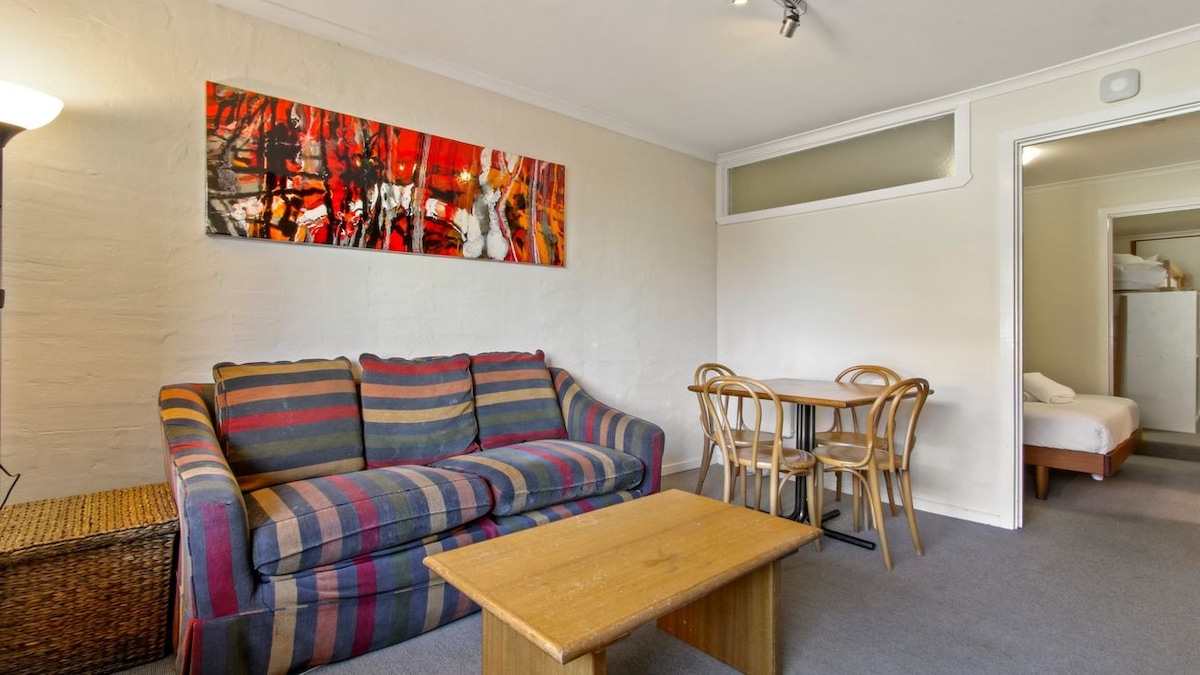 A cozy living space features a striped sofa paired with a wooden coffee table. A simple dining area is visible, accommodating four guests. A vibrant artwork adorns the wall, adding a splash of color to the environment. Soft lighting contributes to a welcoming ambiance.