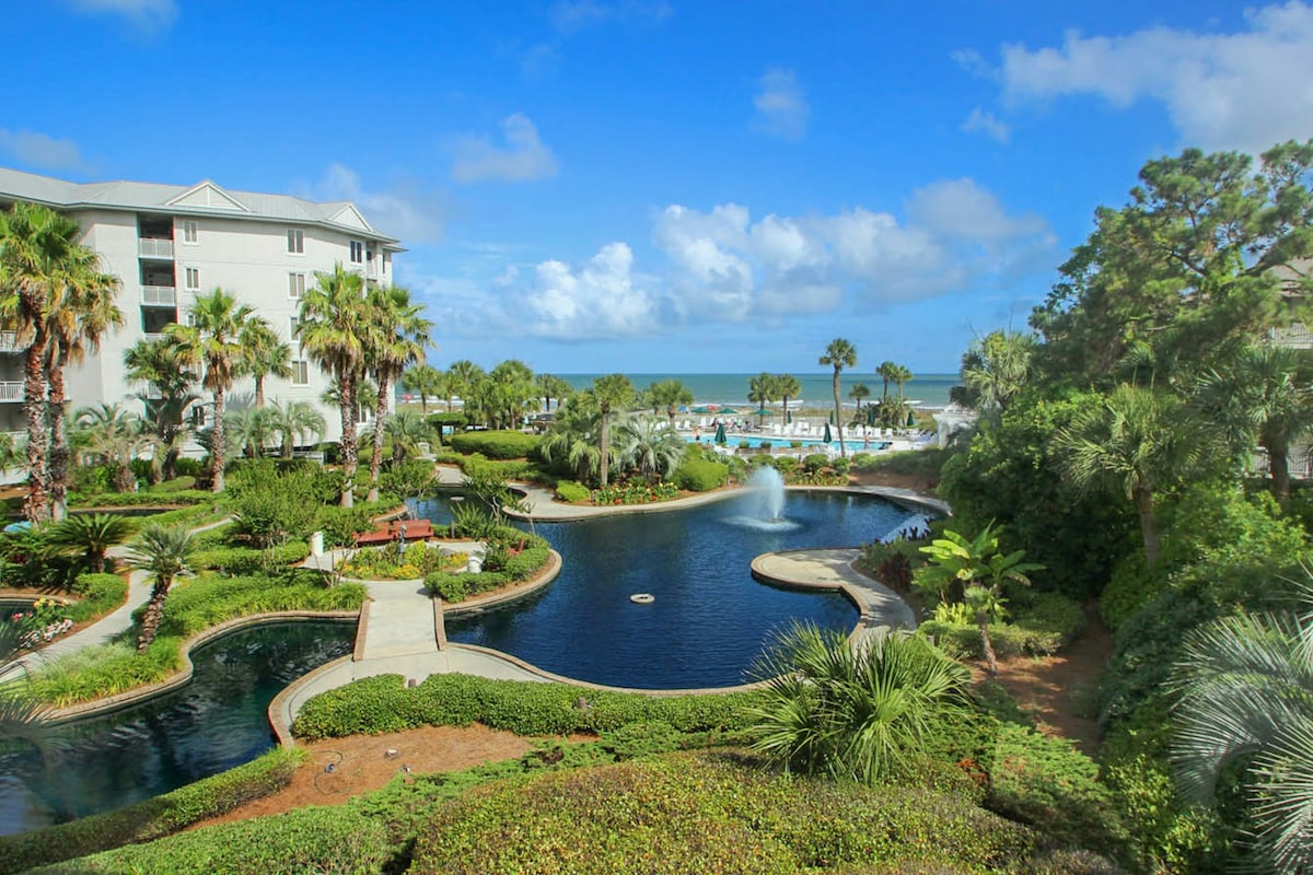 A tranquil view is presented, showcasing lush landscaping with palm trees and verdant plants surrounding a circular lagoon. A fountain is seen in the lagoon, while the beach and ocean are visible in the background under a blue sky with wispy clouds.