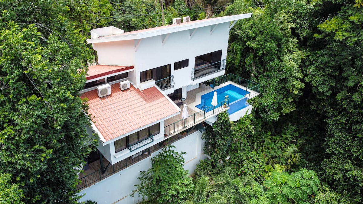 A modern two-level home is nestled in lush greenery, featuring a private pool surrounded by a spacious terrace. Large windows offer a view of the surrounding rainforest and provide natural light to the beautifully designed indoor spaces.