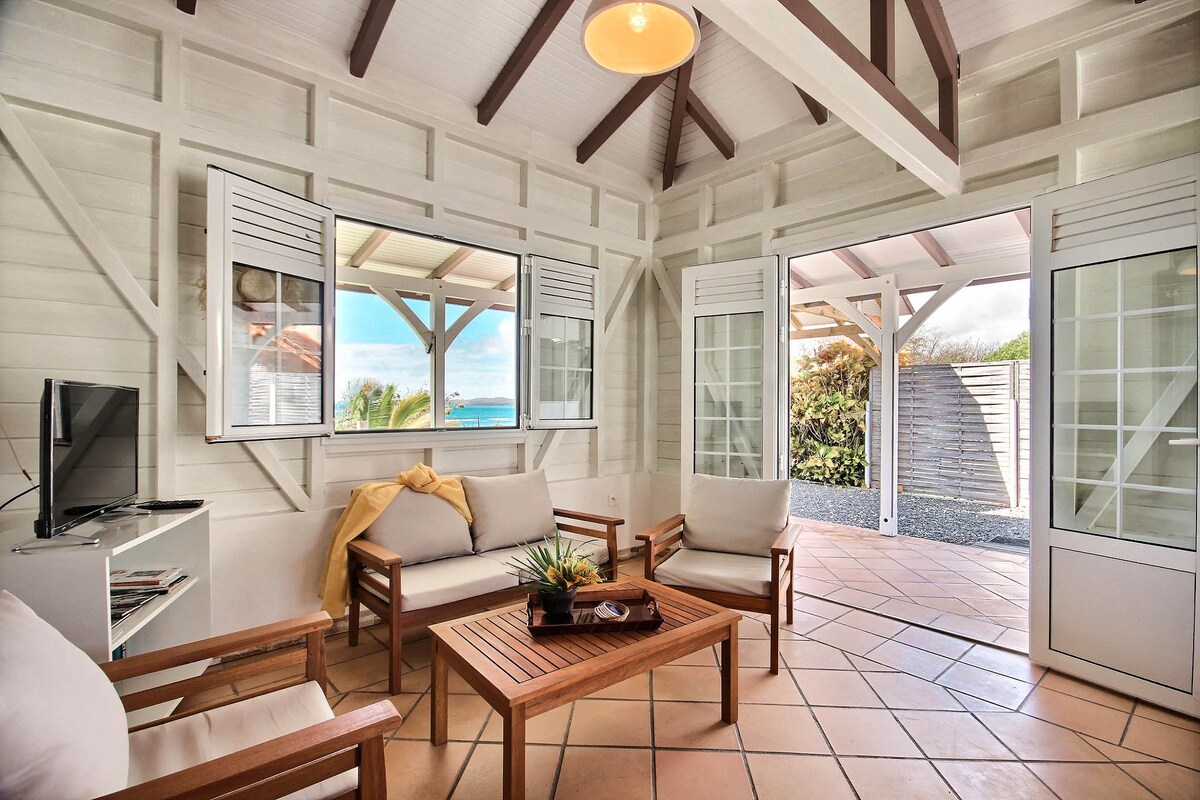 A bright living area features large windows providing natural light and views of the outdoors. Two inviting seating options around a wooden table are arranged, with soft cushions enhancing comfort. A television is positioned nearby, and a warm color palette complements the space.