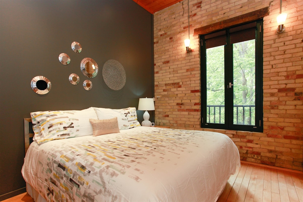 A contemporary bedroom features a spacious bed adorned with a patterned duvet. Reflective circular wall decor adds interest to the dark accent wall. A lamp sits on a nightstand beside the bed. Large windows allow natural light and offer a view of greenery outside.
