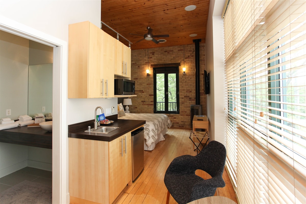 A modern studio space features wooden ceilings and large windows allowing for ample natural light. A kitchenette is equipped with sleek cabinetry and appliances. A cozy sleeping area includes a spacious bed, and additional seating is provided by a stylish chair near the window.