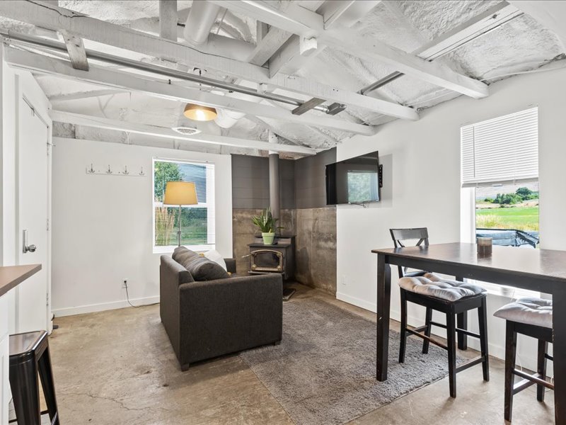 An inviting open living space features a comfortable sofa, a flat-screen TV mounted on the wall, and a small dining table with stools. Natural light filters through the windows, highlighting the minimalist decor and exposed beams on the ceiling.